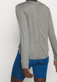 Šedé tričko s dlouhým rukávem a kulatým výstřihem, texturovanou látkou a švem na zádech, doplněné modrými sportovními šortkami.