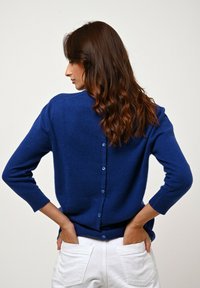 Cardigan di lana blu con chiusura a bottoni sul retro, polsini a costine e una texture liscia. Abbinato a pantaloni bianchi, con una vestibilità morbida.