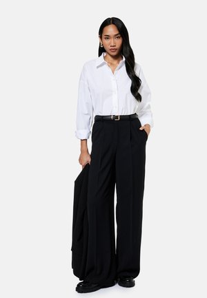 Mujer con cabello largo y negro lleva una camisa blanca abotonada, pantalones negros de pierna ancha con cinturón, sostiene una chaqueta negra, de pie sobre un fondo liso.