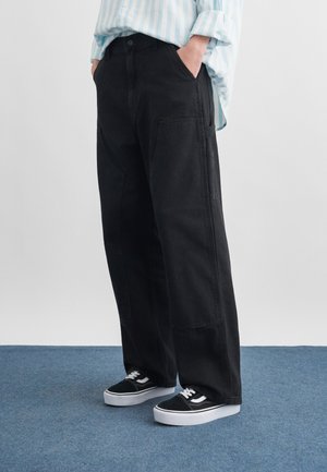 Persoană purtând pantaloni negri largi cu buzunare laterale mari, cu mâinile în buzunare, încălțări sport negre și albe și o cămașă cu dungi albastre deschise.