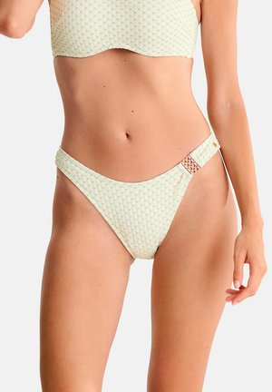 Femme portant un bas de bikini crème texturé avec détail de boucle latérale et haut assorti, montrée de la taille à la cuisse supérieure.