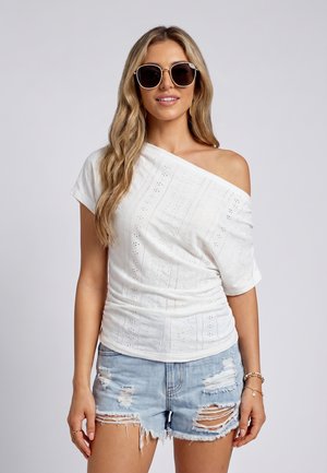 Vrouw met grote zwarte zonnebril, wit off-shoulder eyelet topje en versleten lichte denim shorts, glimlachend tegen een effen achtergrond.