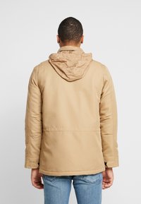 Tan fodrad jacka med huva, med en slät textur och rak skärning, ihop med blå denimjeans.