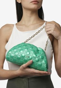 Borsa a tracolla verde trapuntata realizzata in materiale lucido, con una tracolla a catena e un motivo floreale. La borsa è tenuta in mano da una persona.