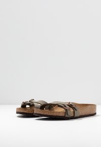 Et par brune korksåler med justerbare olivengrønne stropper og metalspænder på en hvid overflade og ensfarvet baggrund.