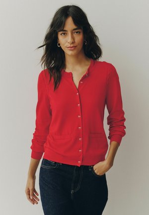 Frau mit dunklen Haaren, die goldene Creolen, eine rote, geknöpfte Strickjacke und dunkle Jeans trägt, steht mit einer Hand in der Tasche.
