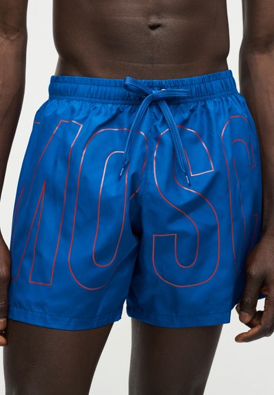 Κοντινό πλάνο ενός ατόμου που φορά μπλε μαγιό βερμούδα με ελαστική ζώνη και κορδόνι, με μεγάλα, περιγραμμικά γράμματα στο ύφασμα.