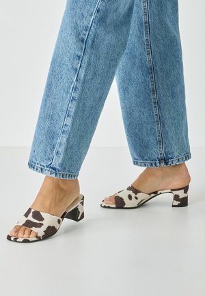 Mules à talons imprimé vache avec une large bride sur le pied, un design à bout ouvert et un talon bloc fin. Le matériau semble doux avec un motif texturé.