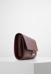 Bordó kožená kabelka s klopou a stříbrným zapínáním, vystavená na bílém podstavci na světle šedém pozadí.