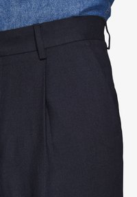Marineblå skræddersyede bukser med en glat tekstur, der har skarpe folder, en flad talje og bæltestropper. En denimskjorte ses delvist ovenfor.