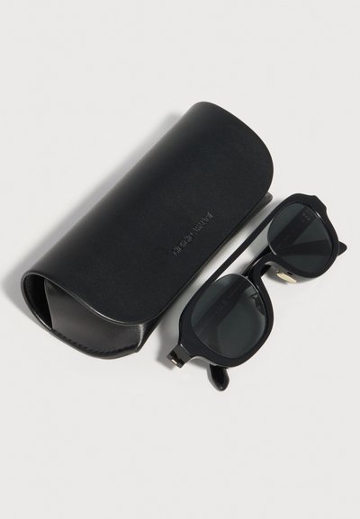 Μαύρα ορθογώνια γυαλιά ηλίου με σκοτεινούς φακούς δίπλα σε μαύρη δερμάτινη θήκη με ανάγλυφη την επιγραφή "Monte Blanc" πάνω σε λευκή επιφάνεια.