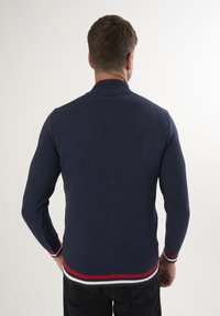 Dunkelblauer gestrickter Pullover mit einem strukturierten Muster, hohem Kragen und kontrastierenden roten und weißen Streifen an den Bündchen und dem Saum. Rückansicht.