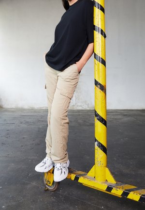 Persoon die beige cargobroek en witte sneakers draagt, leunt tegen een geel-zwarte gestreepte industriële paal, staand op zijn wielen binnen.
