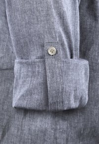 Manga de camisa de lino gris con un detalle de botón en contraste. La tela tiene un acabado texturizado y el puño está cuidadosamente doblado.
