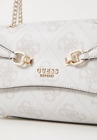 Hvit teksturert håndveske med preget mønster, gullhardware og en kjede-rem. Har merkevarenavnet "GUESS" tydelig vist.