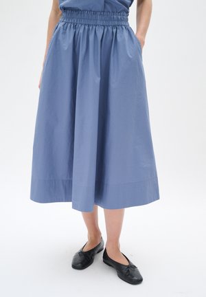 Kobieta ubrana w niebieską midi spódnicę z elastycznym pasem i czarne baleriny, stojąca z rękami w kieszeniach na tle białego tła.