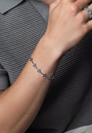 Sølv kædearmbånd med blomstermotiver, teksturerede led og en poleret overflade, båret på et håndled med en subtil tan, mod en grå skjorte.