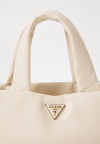 Sac à main en cuir souple couleur crème avec poignée rembourrée et plaque triangulaire dorée du logo Guess USA à l'avant.