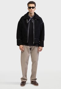 Chaqueta de forro polar negra con detalles de piel, cierre de cremallera y cuello alto, llevada sobre un suéter negro de canalé. Pantalones beige y zapatos marrones completan el conjunto.