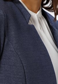 Gros plan sur une personne portant un blazer bleu marine texturé sur un chemisier blanc avec un petit pli près du col.