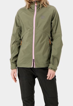 Persoon die een olijfgroene jas met roze voorrits en zwarte broek draagt, staand tegen een effen lichte achtergrond.