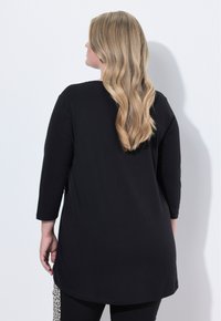 Schwarzes Langarmtop mit abgerundetem Saum, aus weichem Stoff. Sichtbare Seitennaht mit Musterdetail an der Hose. Rückansicht gezeigt.