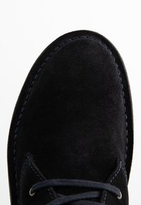 Scarpa in suede nera con punta arrotondata, cuciture blu a contrasto e lacci piatti color navy. Texture morbida con dettagli minimi.