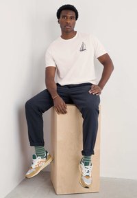 Camiseta de manga corta color rosa claro con un estampado de velero, combinada con pantalones oscuros y zapatillas multicolores, sentado sobre un cubo de madera.