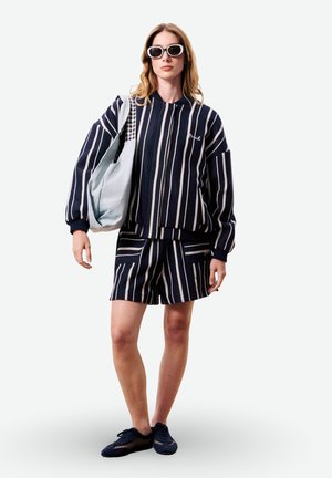 Femme portant une veste et un short à rayures verticales bleu marine et blanches, des baskets en mesh bleu marine, des lunettes de soleil blanches, et portant un sac fourre-tout bleu clair.