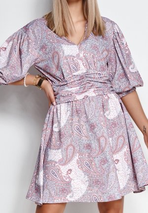 Frau trägt ein Kleid mit rosa-weißem Paisley-Muster und Puffärmeln, dazu Armbänder und eine Uhr als Accessoires.