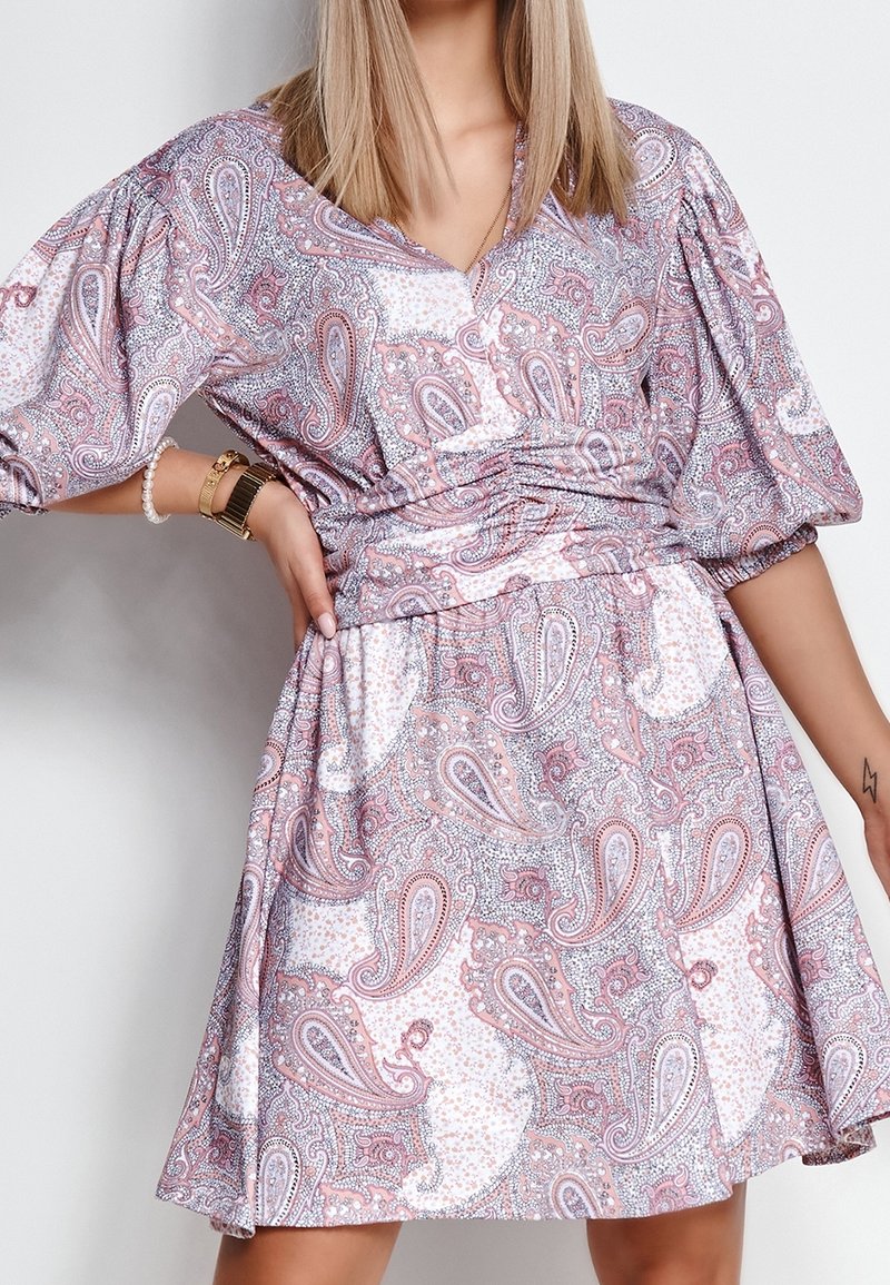 Femme portant une robe à manches bouffantes rose et blanche à motif cachemire, accessoirisée avec des bracelets et une montre.