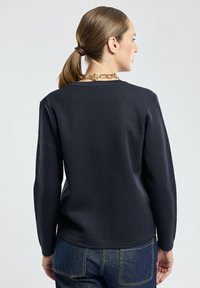 Femme aux cheveux bruns attachés en queue de cheval portant un haut à manches longues noir et un jean bleu, vue de dos sur un fond clair.