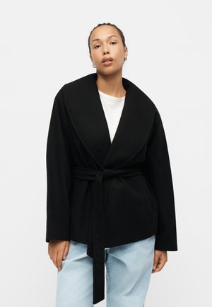 Kvinde iført en sort jakke med bælte over en hvid skjorte og lyseblå jeans, stående mod en ensfarvet lys baggrund.