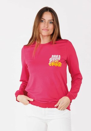 Frau mit langen braunen Haaren, die ein leuchtend pinkes Langarmshirt mit weißem und gelbem Text auf der Brust trägt.