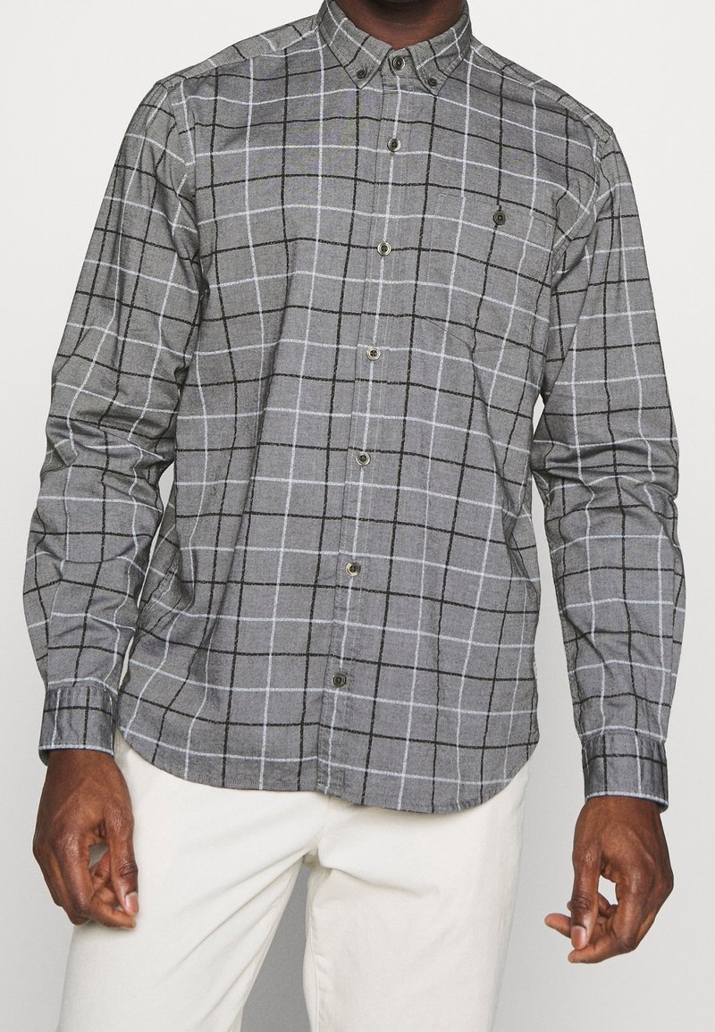 Camicia grigia a quadri con bottoni, con linee a griglia nere e bianche, dotata di una tasca sul petto, colletto strutturato e maniche lunghe. Tessuto testurizzato.