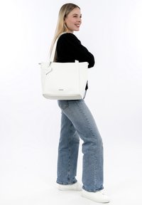 Bolso tote de cuero blanco con forma rectangular, asas superiores y una etiqueta decorativa en forma de corazón. Textura suave con mínima marca.