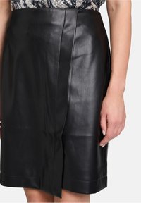 Schwarze Lederrock mit glatter Textur, mit einem vorderen Schlitz, Nahtdetails und geradem Schnitt. Hand zur Größenveranschaulichung gezeigt.