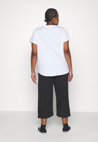 T-shirt blanc à manches courtes et col arrondi, associé à un pantalon noir à jambes larges, se tenant contre un fond uni.
