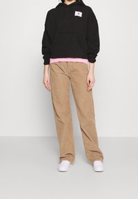Sudadera negra con un bolsillo frontal y un logotipo, superpuesta a una camiseta rosa, combinada con pantalones de pana beige y zapatillas blancas.