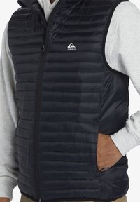 Black padded vest made from glossy material with horizontal quilted sections, zipper front, and side pockets, worn over a grey hoodie.