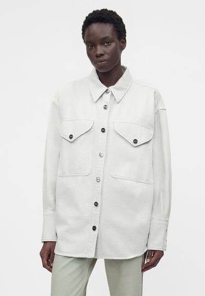 Μαύρη γυναίκα που φοράει λευκό πουκάμισο μεγάλης διάστασης με τσέπες στο στήθος, στέκεται μπροστά σε απλό λευκό φόντο.