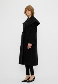 Cappotto di lana nero con ampio collo a scialle, due tasche laterali e silhouette dritta, abbinato a pantaloni neri aderenti e scarpe a tacco basso.
