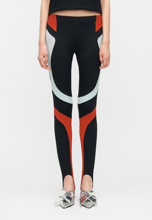 Vista frontale di una persona che indossa leggings neri con motivi geometrici rossi e azzurri, abbinati a scarpe con punte argentate.
