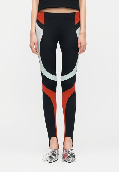 Vue de face d'une personne portant des leggings noirs avec des motifs géométriques rouges et bleu clair, associés à des chaussures argentées à bout pointu.