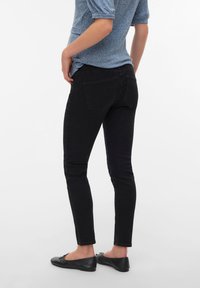 Άτομο που φοράει μαύρο στενό τζιν, μαύρα μπαλέτα και γκριζοπράσινο κοντομάνικο τοπ, στέκεται μπροστά σε λευκό φόντο.