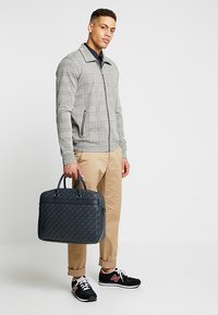 Veste zippée à carreaux gris, pantalon kaki, baskets noires. Tient une mallette texturée bleu foncé avec poignées et poche zippée à l'avant.