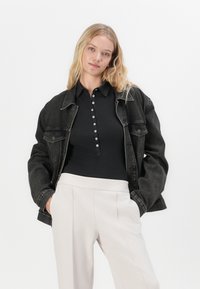 Zwarte denimjack over een zwarte geribde polo met witte knopen, gecombineerd met lichtbeige hoge taillebroeken met zijzakken.