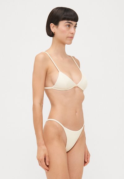 Crèmekleurige driehoekige bikini met ribbeltextuur, dunne verstelbare bandjes en bijpassende low-rise broek. Eenvoudige scalloped randdetails.