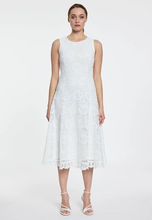 Vrouw staand, gekleed in een mouwloze witte kanten midi-jurk met bloemmotieven en witte sandalen met bandjes en hoge hakken tegen een effen achtergrond.