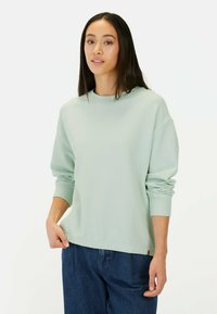 Mintgroene sweatshirt met textuur, ronde hals, losse pasvorm en lange mouwen, gecombineerd met donkere spijkerbroeken. Subtiele branding zichtbaar.